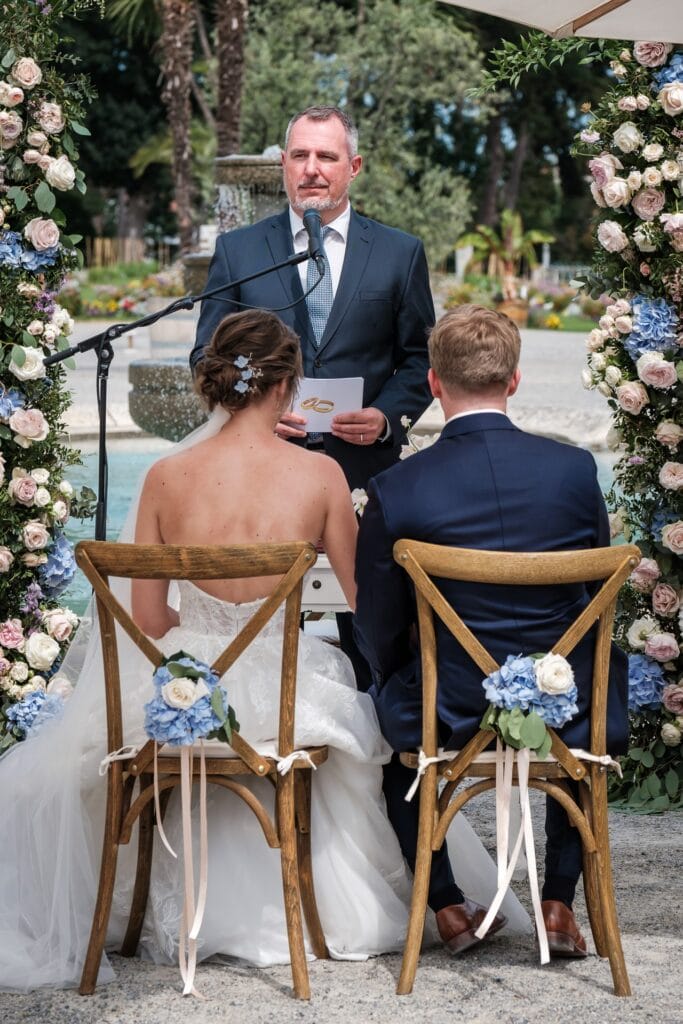 Hochzeitskrone Ein Paar sitzt während einer Hochzeitszeremonie im Freien mit dem Rücken zur Kamera. Ein Mann steht am Mikrofon und spricht inmitten wunderschöner Blumenarrangements – die Essenz authentischer Hochzeitsfotos eines talentierten Hochzeitsfotografen aus Wien und Niederösterreich.