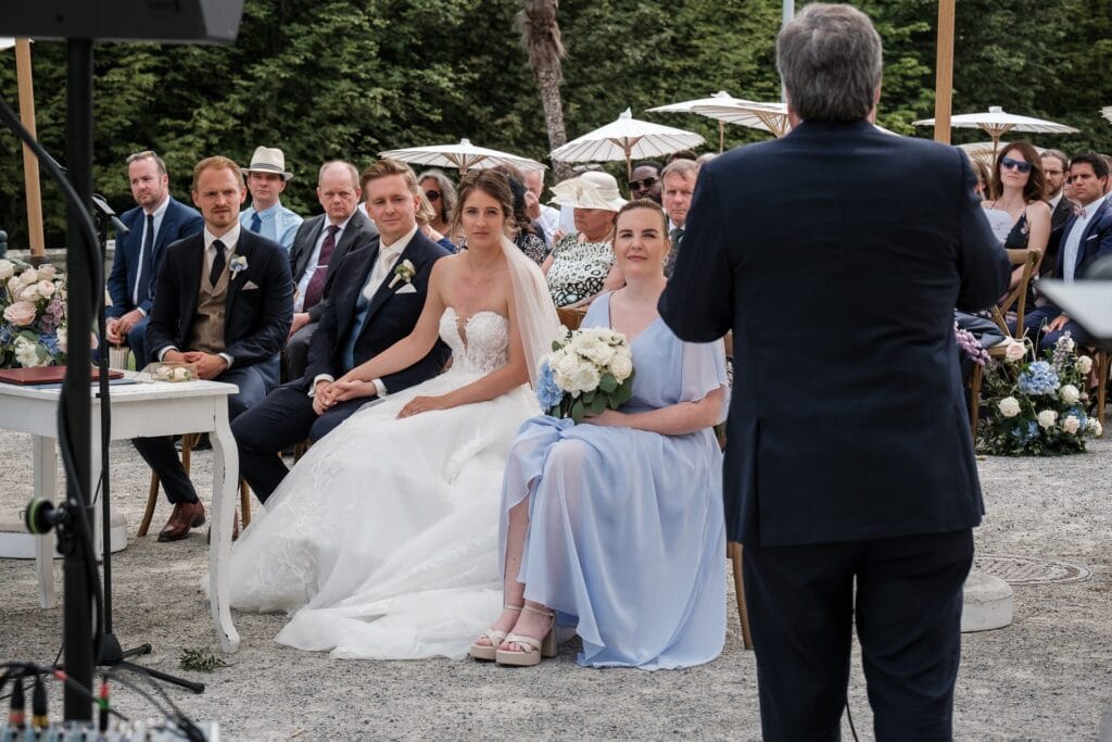 Hochzeitskrone Hochzeitszeremonie im Freien mit einer Braut im weißen Kleid und einem Bräutigam im Anzug, die zwischen den Gästen sitzen. Ein Mann im Anzug hält eine Rede vorn. Der Hochzeitsfotograf in Wien und Niederösterreich fängt echte Momente ein und sorgt für authentische Fotos, während die Gäste Sonnenschirme zwischen blühenden Blumen halten.