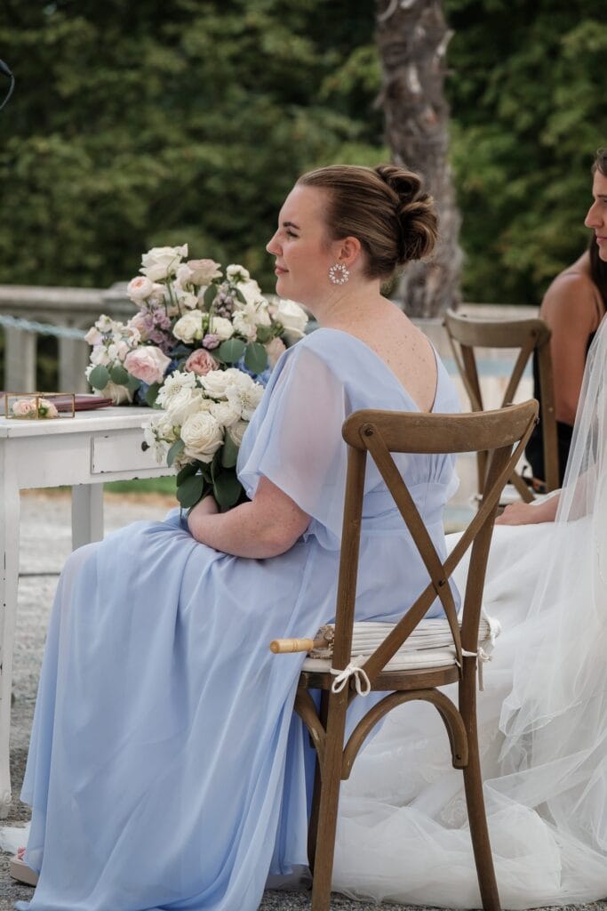 Hochzeitskrone Eine Frau in einem hellblauen Kleid hält einen Blumenstrauß und fängt echte Momente ein, während sie im Freien auf einem Holzstuhl sitzt, was an den Stil erinnert, den ein authentisches Hochzeitsfoto anstreben würde.