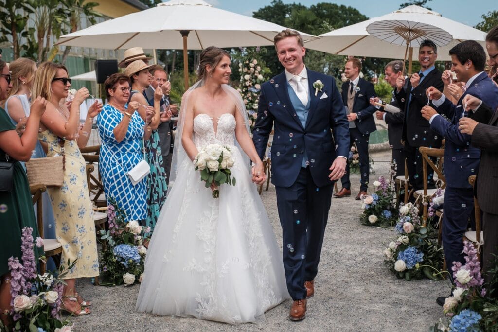 Hochzeitskrone Braut und Bräutigam schreiten draußen den Gang entlang, umgeben von Gästen, die sie mit Konfetti überschütten. Sie trägt ein weißes Kleid und hält Blumen in der Hand, er einen dunklen Anzug. Die Gäste halten Regenschirme in der Hand und halten echte Momente fest, die zu natürlichen Hochzeitsfotos werden, die für immer in Erinnerung bleiben.