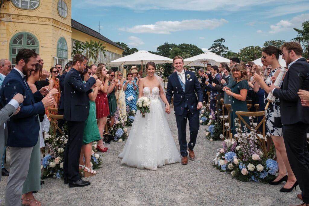 Hochzeitskrone Das Brautpaar strahlt vor Freude und schreitet Hand in Hand den Gang im Freien entlang, umgeben von jubelnden Gästen. Diese echten Momente werden wunderschön als natürliche Erinnerungen festgehalten, die man für immer in Ehren halten wird.