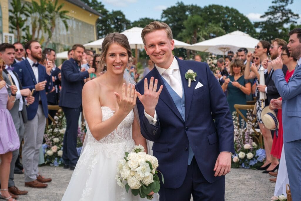 Hochzeitskrone Eine Braut und ein Bräutigam in Hochzeitskleidung zeigen stolz ihre Ringe in die Kamera, umgeben von Gästen im Freien. Aufgenommen von einem Hochzeitsfotografen in Wien und Niederösterreich, offenbaren diese natürlichen Hochzeitsfotos nur echte Momente der Freude und Liebe.