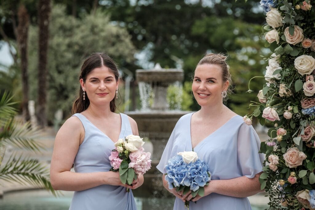 Hochzeitskrone Zwei Frauen in hellblauen Kleidern, Blumensträuße in den Händen, stehen vor einem Brunnen neben einem Blumenbogen, festgehalten mit dem kunstvollen Auge eines Hochzeitsfotografen in Wien und Niederösterreich. Dieser Moment verkörpert natürliche Hochzeitsfotos vom Feinsten.