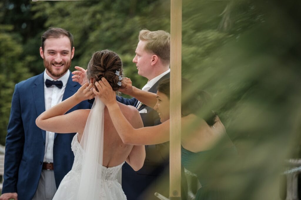 Hochzeitskrone Eine Frau rückt im Freien den Schleier einer Braut zurecht und macht authentische Hochzeitsfotos, während zwei Männer in der Nähe lächeln und diese echten Momente genießen.