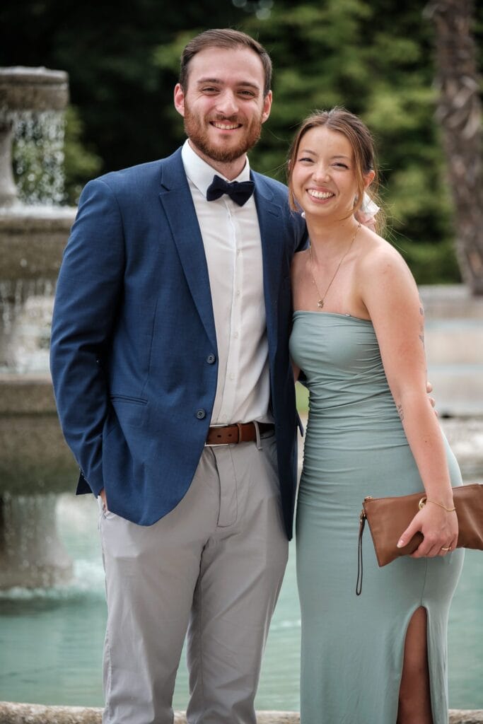 Hochzeitskrone Ein Mann in einem blauen Blazer und einer Fliege steht neben einer Frau in einem trägerlosen grünen Kleid. Beide lächeln strahlend vor einem Springbrunnen im Freien, festgehalten in einem jener echten Momente, die wirklich authentische Hochzeitsfotos ausmachen.