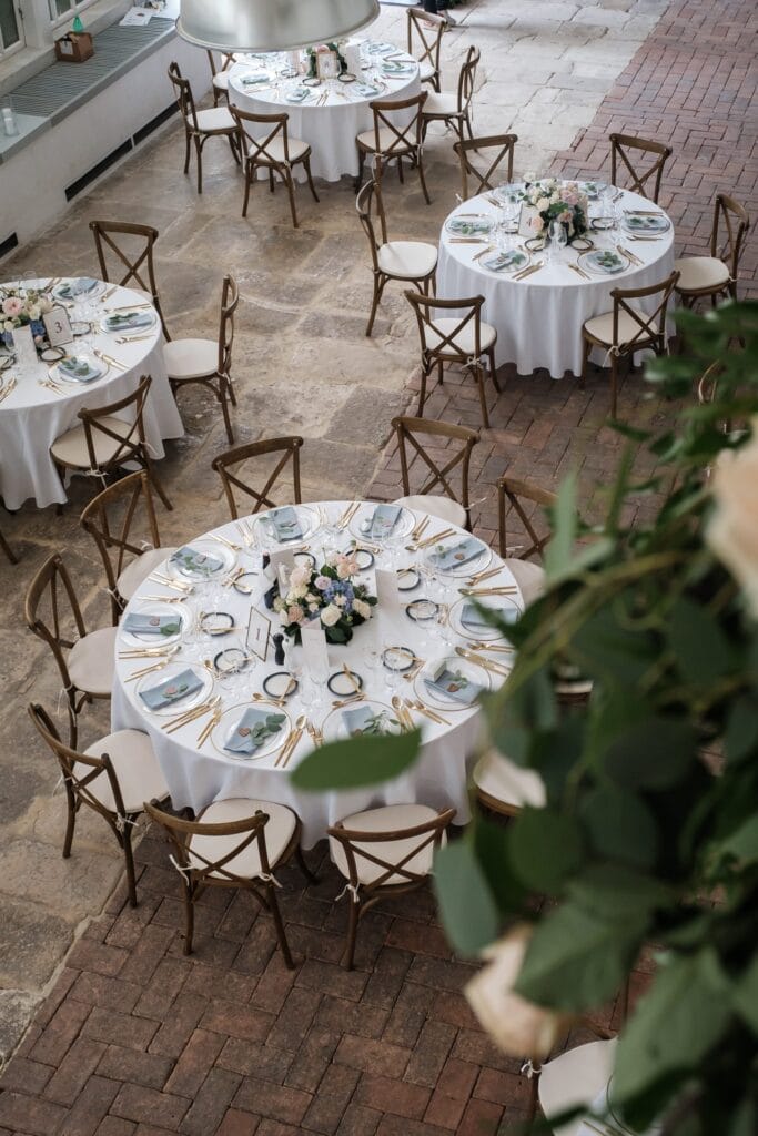 Hochzeitskrone Elegante Hochzeitstische mit weißen Tischdecken, Blumengestecken und hübsch angeordnetem Geschirr; Holzstühle umgeben jeden Tisch auf einem Boden aus Stein und Ziegeln, perfekt für die Aufnahme authentischer Hochzeitsfotos, die echte Momente der Feier zeigen.