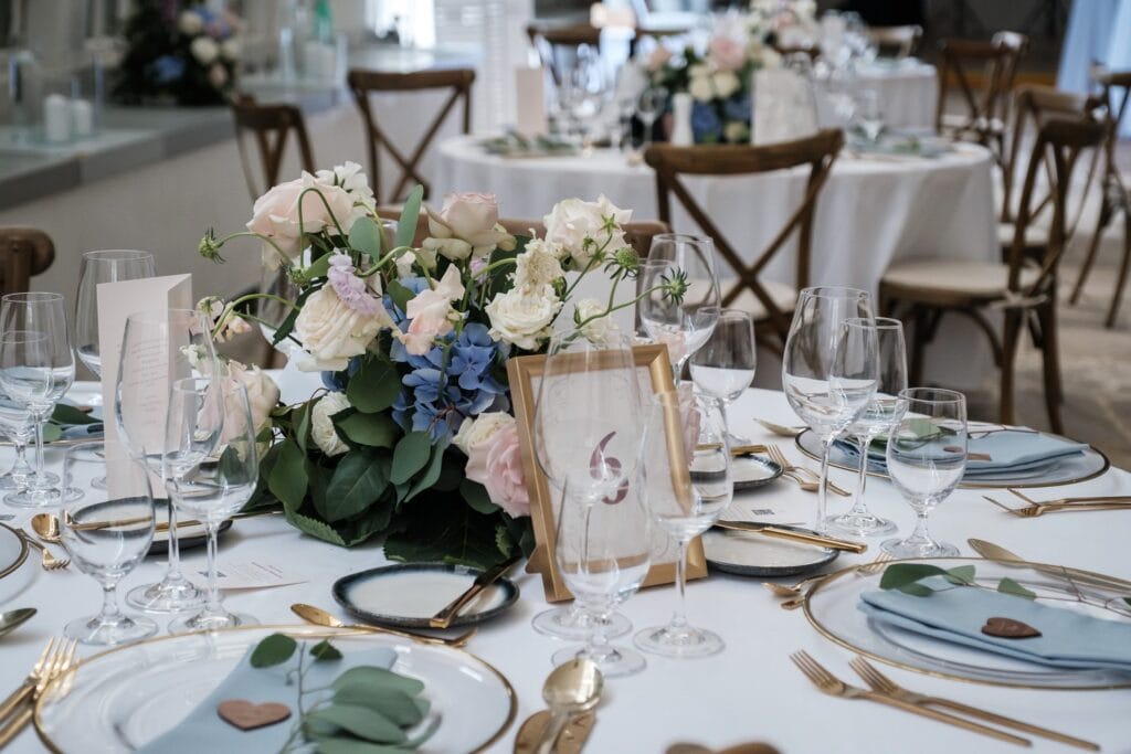 Hochzeitskrone Elegante Tischdekoration für den Hochzeitsempfang mit Blumenschmuck, goldenem Besteck, Gläsern, hellblauen Servietten und einer gerahmten Tischnummer – fängt echte Momente für alle ein, die nach authentischen Hochzeitsfotos suchen.