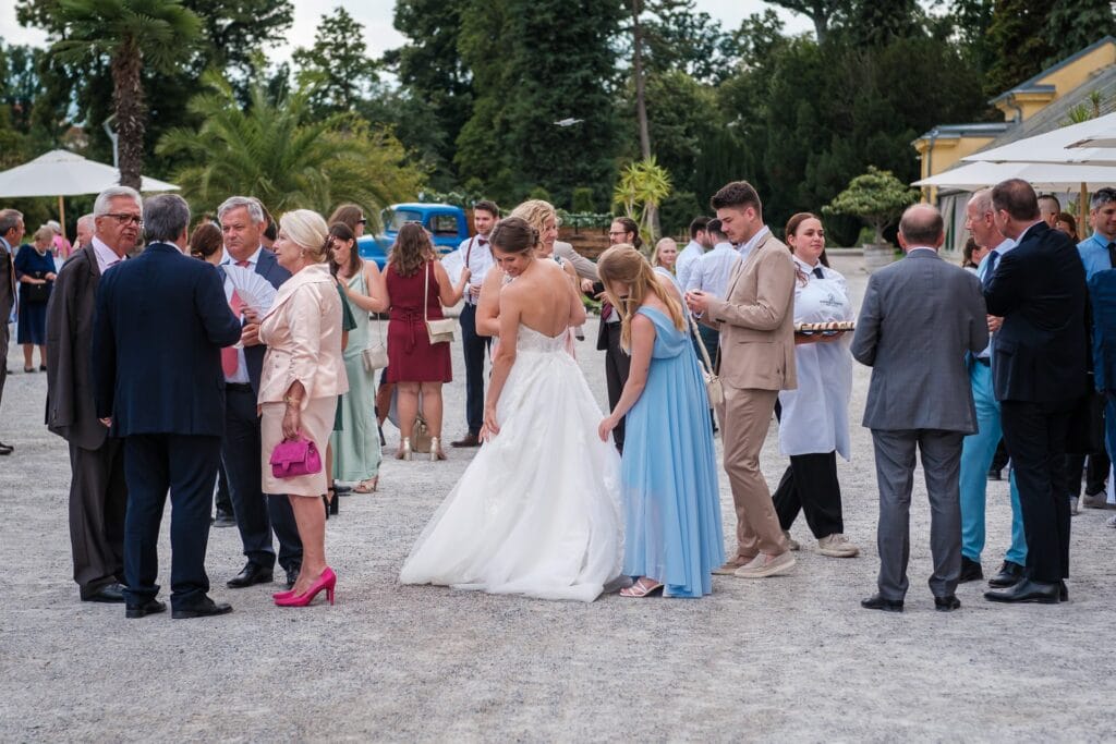 Hochzeitskrone Eine Gruppe von Menschen in formeller Kleidung versammelt sich im Freien bei einer Veranstaltung. Die Kulisse umfasst Palmen und einen Kiesweg. In der Mitte steht eine Frau im Hochzeitskleid, während ein Hochzeitsfotograf in Wien und Niederösterreich authentische Hochzeitsfotos dieser echten Momente einfängt.