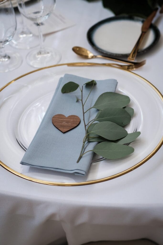 Hochzeitskrone Auf einem weißen Teller mit Goldrand liegen eine blaue Serviette, ein Herz aus Holz und ein Zweig mit Blättern. Eine silberne Gabel und ein silberner Löffel ergänzen die Szene auf einer weißen Tischdecke und fangen echte Momente ein, die an natürliche Hochzeitsfotos eines erfahrenen Hochzeitsfotografen in Wien und Niederösterreich erinnern.