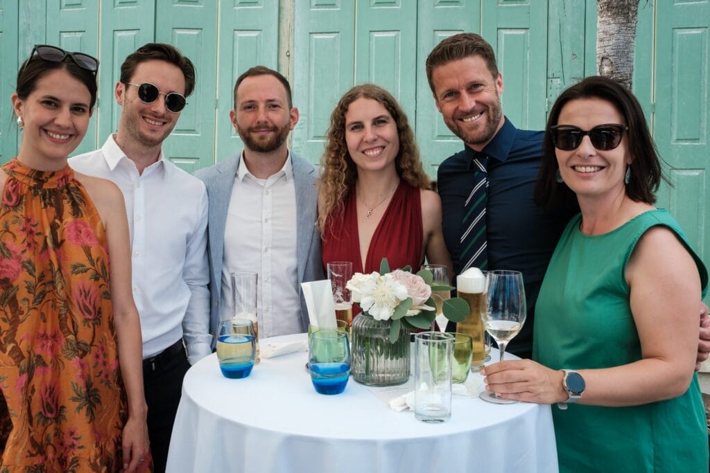 Hochzeitskrone Sechs Personen stehen um einen mit Getränken und Blumen geschmückten Tisch und fangen echte Lächeln für ein Foto vor dem Hintergrund blaugrüner Türen ein und zeigen „echte Momente in natürlichen Hochzeitsfotos“.