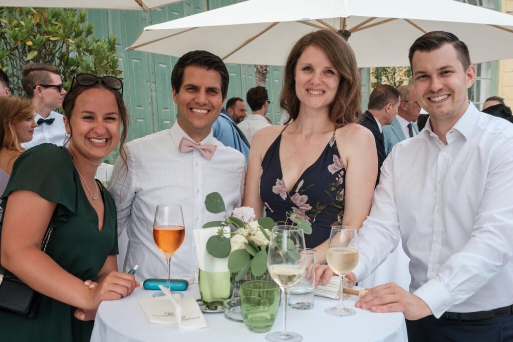 Hochzeitskrone Vier Personen stehen lächelnd um einen Tisch bei einer Veranstaltung im Freien, mit Weingläsern und einem Blumenschmuck in der Mitte, und fangen echte Momente ein. Im Hintergrund verleihen Sonnenschirme und andere Gäste der Szene einen besonderen Charme, der an natürliche Hochzeitsfotos eines Hochzeitsfotografen in Wien und Niederösterreich erinnert.