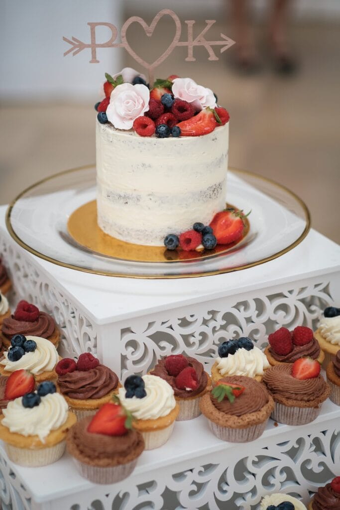 Hochzeitskrone Auf einem weißen Dekoständer steht ein zweistöckiger Kuchen mit Beeren und Blumen darauf, umgeben von verschiedenen Cupcakes mit Zuckerguss und Beeren. Halten Sie diese echten Momente mit authentischen Hochzeitsfotos von einem erfahrenen Hochzeitsfotografen in Wien und Niederösterreich fest.