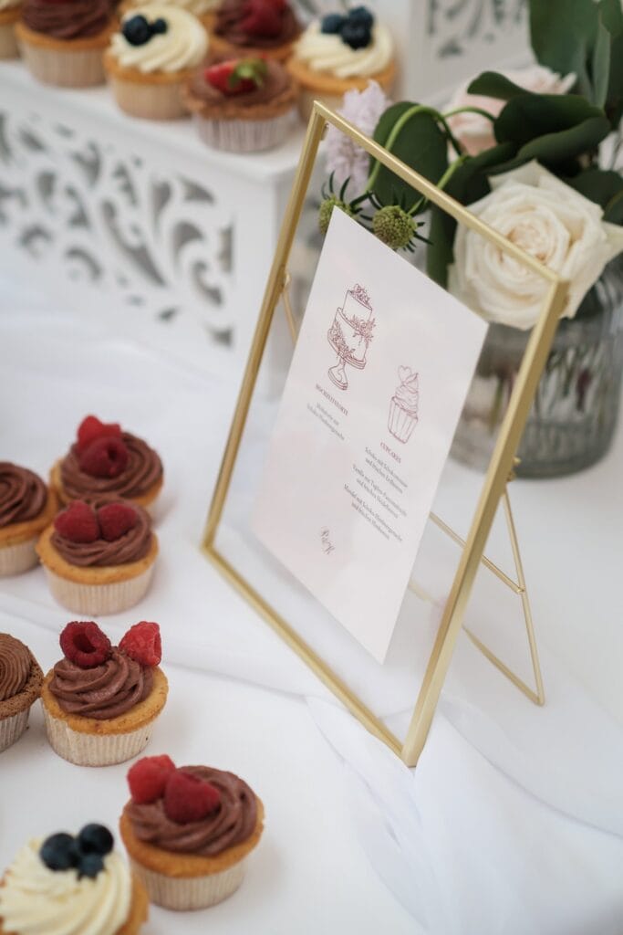 Hochzeitskrone Cupcakes mit Schokoladen- und Vanilleglasur auf einem Tisch neben einem Menü in einem goldenen Rahmen erwecken den Charme authentischer Hochzeitsfotos. Eine Vase mit weißen Blumen ziert den Hintergrund und fängt einen Moment ein, der so zeitlos ist, wie ihn jeder Hochzeitsfotograf in Wien und Niederösterreich einfangen könnte.