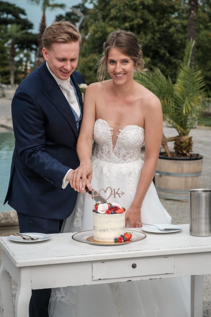 Hochzeitskrone Ein Paar in festlicher Kleidung schneidet eine kleine Hochzeitstorte an, die mit Beeren und Blumen verziert ist. Sie stehen hinter einem weißen Tisch im Freien, umgeben von üppigem Grün, und fangen echte Momente ein, die an authentische Hochzeitsfotos erinnern, die von einem Hochzeitsfotografen in Wien und Niederösterreich aufgenommen wurden.