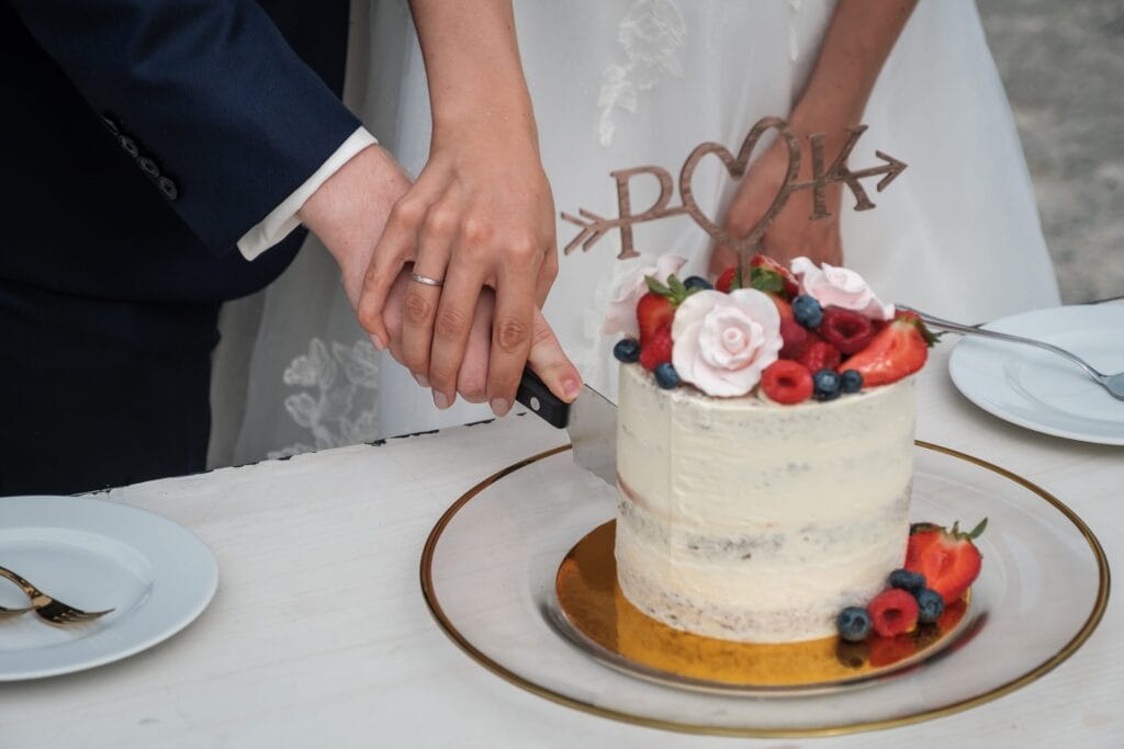 Hochzeitskrone Braut und Bräutigam schneiden eine mit Beeren und Blumen geschmückte, weiß glasierte Torte an, während sie in authentischen Hochzeitsfotos festgehalten werden. Der dekorative Aufsatz ist mit verschlungenen Initialen verziert und hält echte Momente ihres besonderen Tages fest.