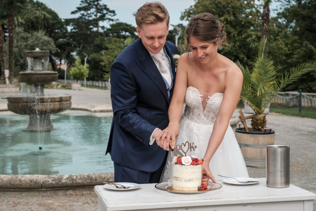 Hochzeitskrone Braut und Bräutigam teilen in Abendgarderobe authentische Hochzeitsmomente, während sie im Freien neben einem Brunnen eine kleine Torte anschneiden und natürliche Hochzeitsfotos schießen, die ihre wahre Liebesgeschichte erzählen.