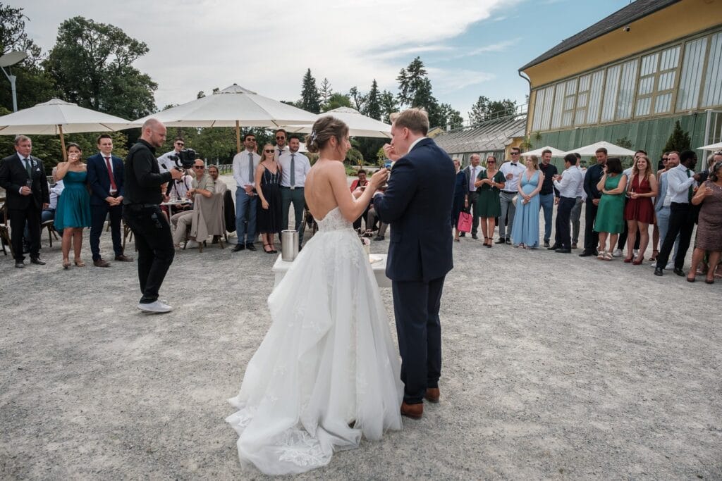 Hochzeitskrone In einem bezaubernden Innenhof stehen Braut und Bräutigam Seite an Seite, umgeben von Gästen bei ihrem Empfang im Freien. Die Szene wird von einem Hochzeitsfotografen in Wien und Niederösterreich wunderschön eingefangen und bietet authentische und natürliche Hochzeitsfotos, die für immer in Erinnerung bleiben.