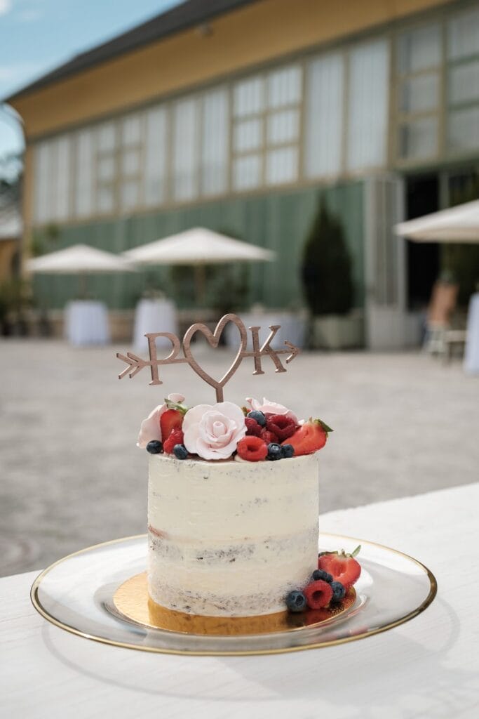 Hochzeitskrone Ein kleiner, mit Zuckerguss überzogener Kuchen, verziert mit Beeren und einem „P <3 K“-Aufsatz, steht auf einer Glasplatte. Diese bezaubernde Szene fängt die Essenz authentischer Hochzeitsmomente in der Nähe eines Gebäudes mit großen Fenstern und Sonnenschirmen ein, perfekt für jeden Hochzeitsfotografen in Wien und Niederösterreich.