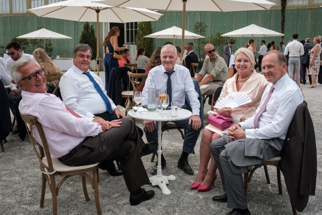 Hochzeitskrone Unter weißen Sonnenschirmen versammeln sich fünf elegant gekleidete Personen mit Getränken um einen Tisch im Freien, umgeben von üppigem Grün. Das Einfangen dieser echten Momente erweckt die Szene zum Leben und erinnert an die Arbeit, die man von einem Hochzeitsfotografen in Wien und Niederösterreich erwartet.