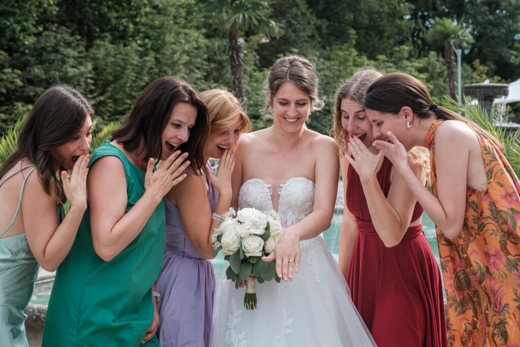 Hochzeitskrone Braut zeigt sechs Brautjungfern im Freien ihren Ring, alle gefangen in echten Momenten der Aufregung, wodurch authentische Hochzeitsfotos entstehen.