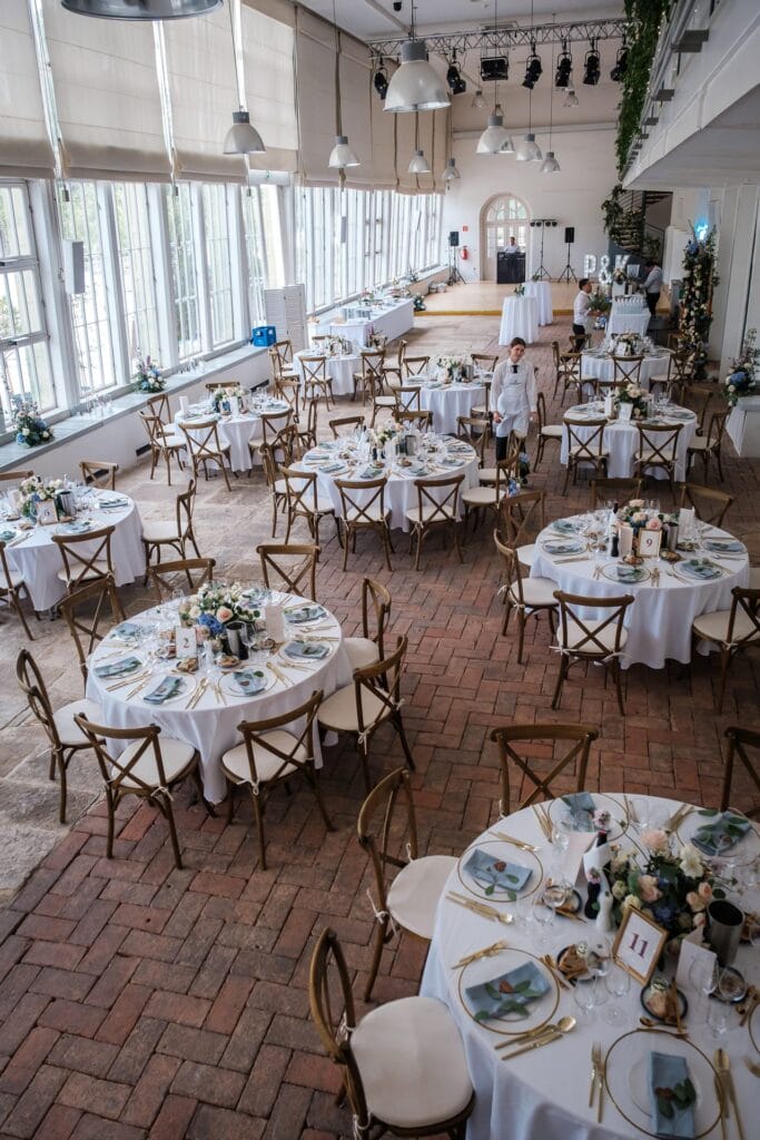 Hochzeitskrone Esstische mit weißen Tischdecken und Blumenschmuck sind in einem geräumigen, gut beleuchteten Veranstaltungsort arrangiert. Eine Person deckt die Tische ab und fängt echte Momente für natürliche, unverfälschte Erinnerungen ein, ganz wie es ein Hochzeitsfotograf in Wien und Niederösterreich tun würde.