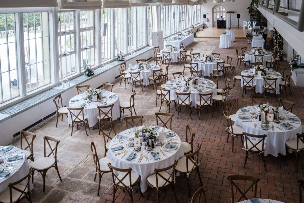 Hochzeitskrone Ein Hochzeitsempfang mit runden, weiß gedeckten Tischen, gedeckt für das Essen in einem sonnendurchfluteten Raum mit großen Fenstern und Holzstühlen, perfekt für die Aufnahme natürlicher Hochzeitsfotos, die echte Momente hervorheben.