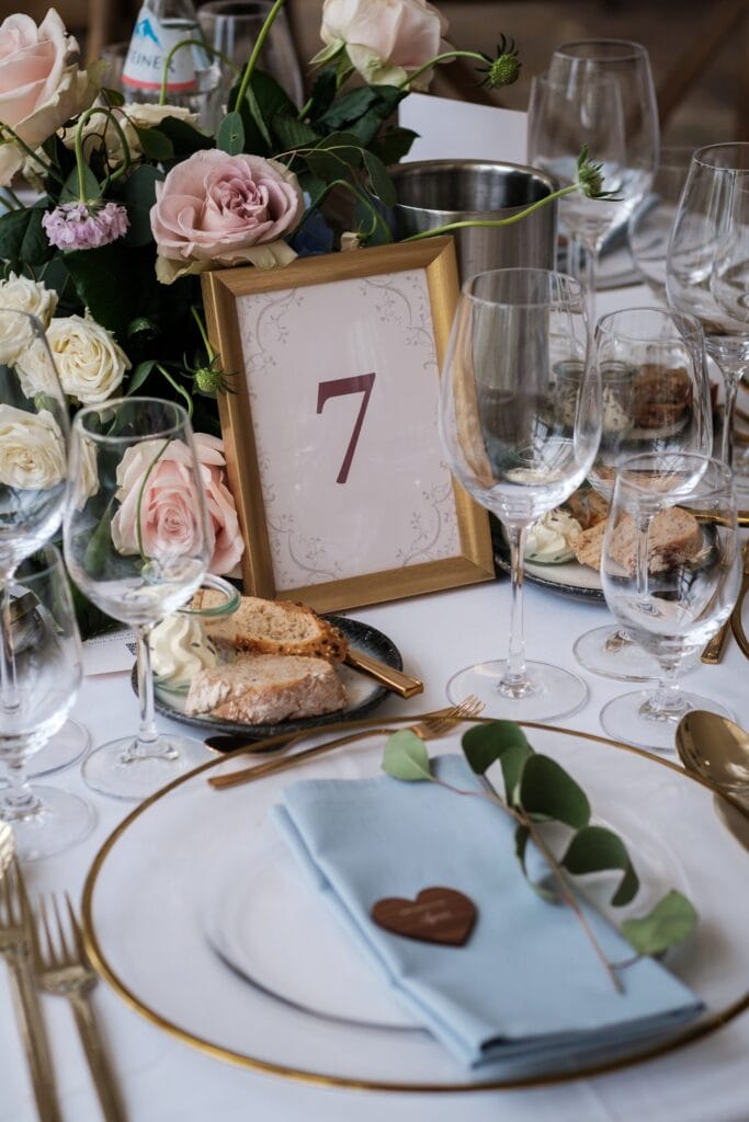 Hochzeitskrone Elegantes Tischgedeck mit floralem Mittelstück, gerahmter Tischnummer 7, Weingläsern, Tellern mit blauen Servietten und einem Schokoladenherz – perfekt für die Aufnahme authentischer Hochzeitsfotos von einem talentierten Hochzeitsfotografen in Wien und Niederösterreich.