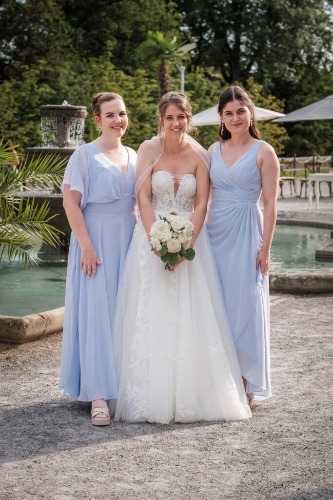 Hochzeitskrone Die Braut in einem weißen Kleid mit Blumen in der Hand steht neben einem Brunnen und wird von zwei Brautjungfern in hellblauen Kleidern flankiert. Diese natürlichen Hochzeitsfotos wurden von einem Hochzeitsfotografen in Wien und Niederösterreich aufgenommen und spiegeln echte Momente perfekt wider.