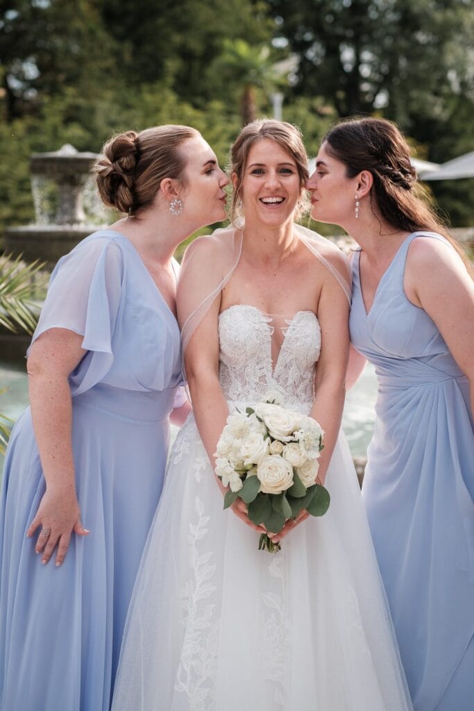 Hochzeitskrone Die Braut lächelt und hält Blumen, während zwei Brautjungfern in hellblauen Kleidern sie im Freien auf die Wangen küssen, eingefangen in „echte Momente“.