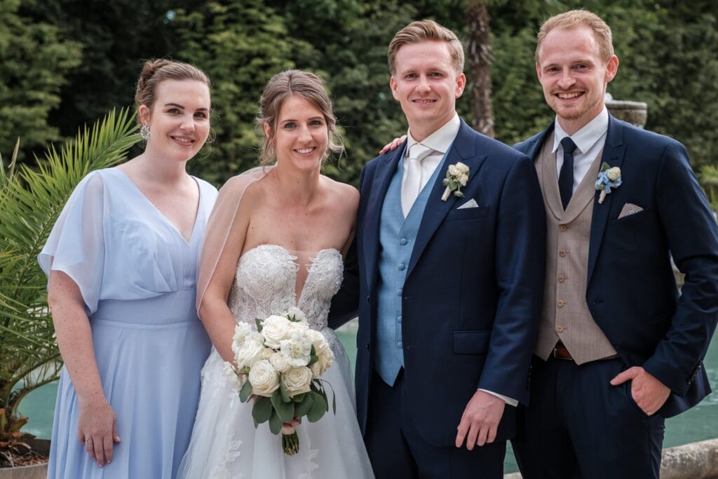 Hochzeitskrone Eine Braut in einem weißen Kleid mit einem Blumenstrauß steht neben einem Bräutigam im blauen Anzug und zwei Freundinnen in eleganter Kleidung. Eingefangen von einem Hochzeitsfotografen in Wien und Niederösterreich, ist diese Szene der Inbegriff authentischer Hochzeitsfotos voller echter Momente.