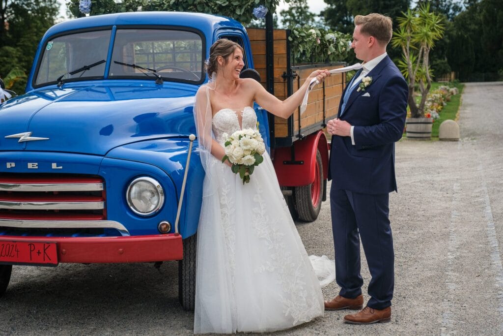 Hochzeitskrone Eine Braut deutet spielerisch auf einen Bräutigam vor einem blauen Oldtimer-Lastwagen und fängt so echte Momente ein. Sie hält einen Blumenstrauß in der Hand und beide tragen Hochzeitskleidung. So entstehen authentische Hochzeitsfotos, die jeder Hochzeitsfotograf in Wien und Niederösterreich bewundern würde.