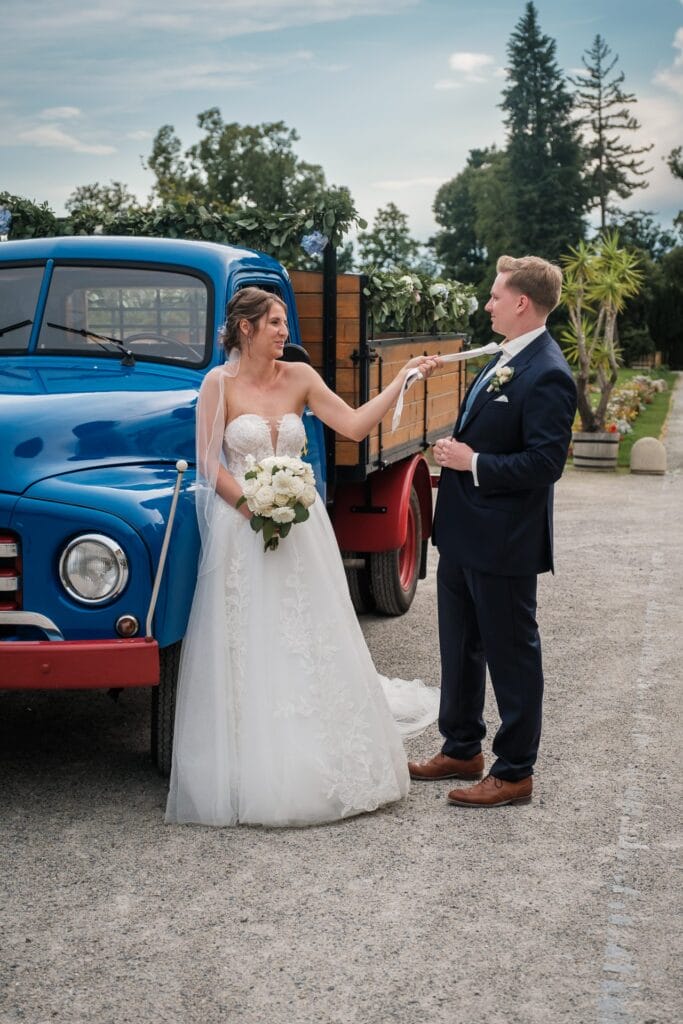 Hochzeitskrone Braut und Bräutigam stehen neben einem blauen Oldtimer-Lastwagen, festgehalten in echten Momenten. Die Braut hält spielerisch lächelnd die Krawatte des Bräutigams. Sie trägt ein weißes Kleid; er trägt einen dunklen Anzug. Perfekt für alle, die natürliche Hochzeitsfotos mit einem Hochzeitsfotografen in Wien und Niederösterreich suchen.