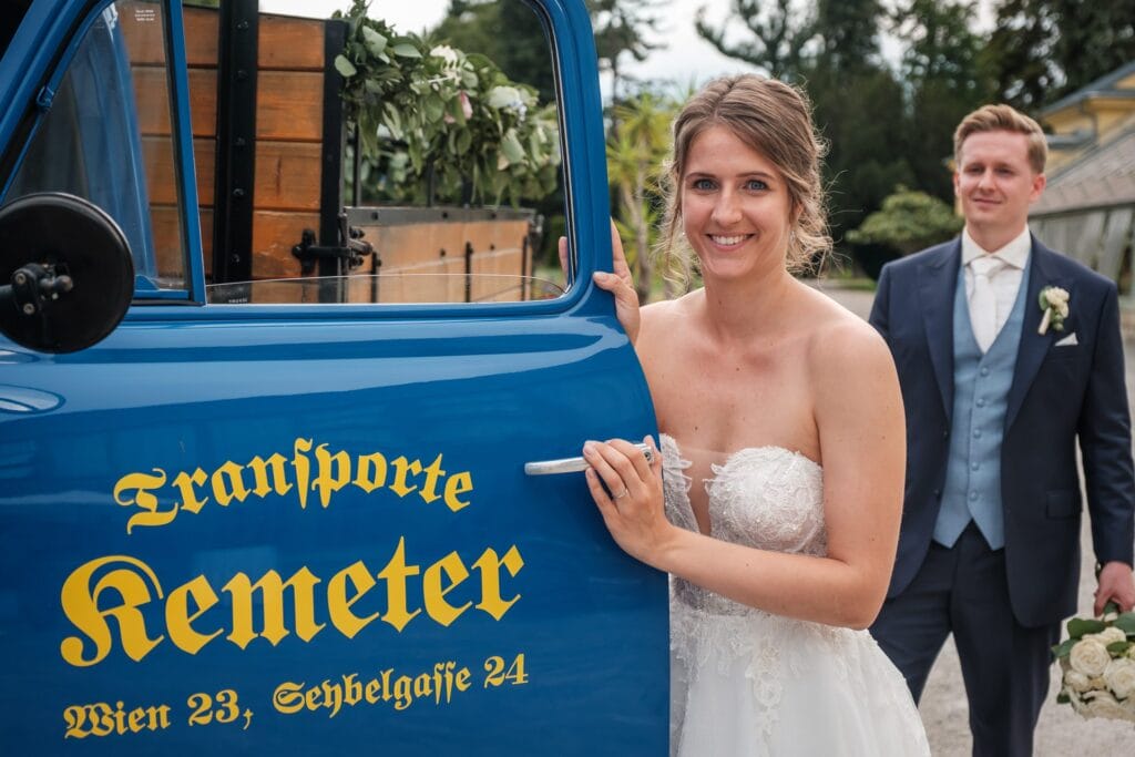 Hochzeitskrone In einer bezaubernden Szene steht die Braut in ihrem weißen Kleid neben einem blauen Oldtimer mit der Aufschrift „Transporte Kemeter“. In der Nähe schaut der Bräutigam zu. Umgeben von Bäumen und Gebäuden ist es eine perfekte Kulisse für authentische Hochzeitsfotos, um echte Momente mit einem Hochzeitsfotografen in Wien und Niederösterreich festzuhalten.