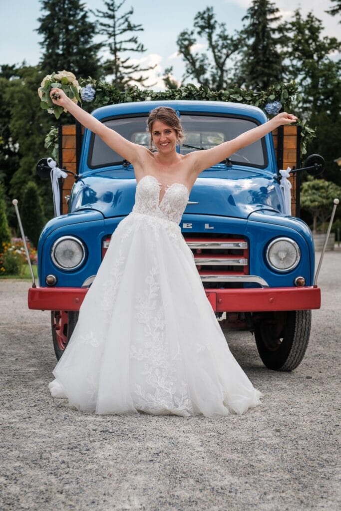 Hochzeitskrone Eine Braut in einem weißen Hochzeitskleid steht mit erhobenen Armen vor einem mit Blumen geschmückten blauen Oldtimer-Truck und fängt echte Momente ein, die nur ein Hochzeitsfotograf in Wien und Niederösterreich durch natürliche Hochzeitsfotos einfangen kann.