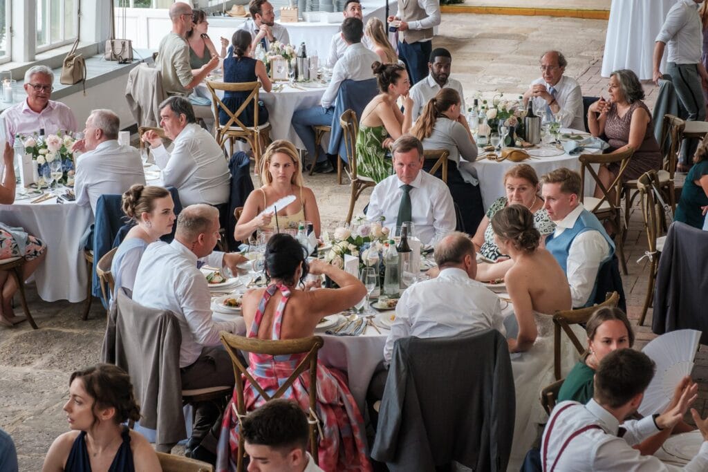 Hochzeitskrone Eine Gruppe von Menschen sitzt an runden Tischen mit weißen Tischdecken, isst und unterhält sich bei einer formellen Veranstaltung im Innenbereich. Blumen und Weinflaschen schmücken die Tische, während ein Hochzeitsfotograf in Wien und Niederösterreich diese echten Momente in natürlichen Hochzeitsfotos einfängt.