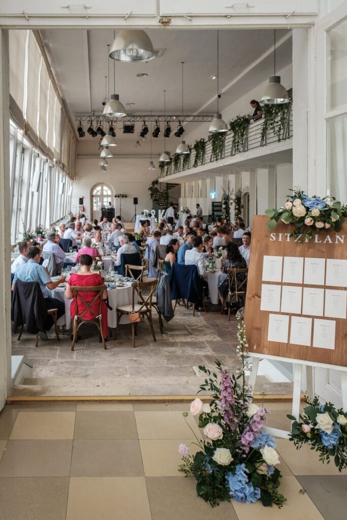 Hochzeitskrone Eine Hochzeitsfeier in einem hellen Raum mit langen Tischen und sitzenden Gästen fängt echte Momente ein. Ein Sitzplan auf einer mit Blumen geschmückten Holztafel begrüßt die Gäste am Eingang.