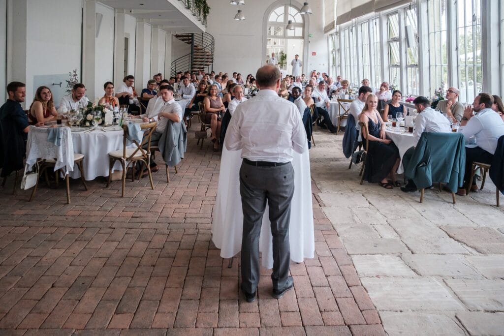 Hochzeitskrone Ein Mann steht an einem Podium und spricht vor sitzenden Gästen und fängt echte Momente einer formellen Veranstaltung in einem hellen Raum mit großen Fenstern und Ziegelboden ein.