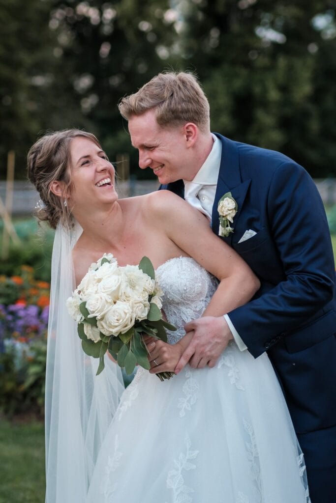 Hochzeitskrone Braut und Bräutigam lachen, die Braut hält weiße Rosen, draußen in festlicher Kleidung mit üppigem Grün. Diese authentischen Hochzeitsfotos fangen echte Momente ein und zeigen wunderschön ihre Freude und Liebe.