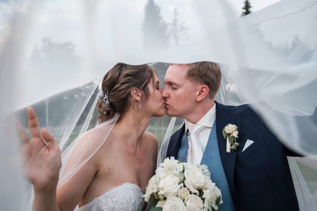 Hochzeitskrone Braut und Bräutigam küssen sich unter dem Schleier und halten echte Momente fest; Die Braut hält einen Strauß weißer Rosen in der Hand, beide in Hochzeitskleidung. Perfekt für alle, die natürliche Hochzeitsfotos suchen, ideal für jeden Hochzeitsfotografen in Wien und Niederösterreich.