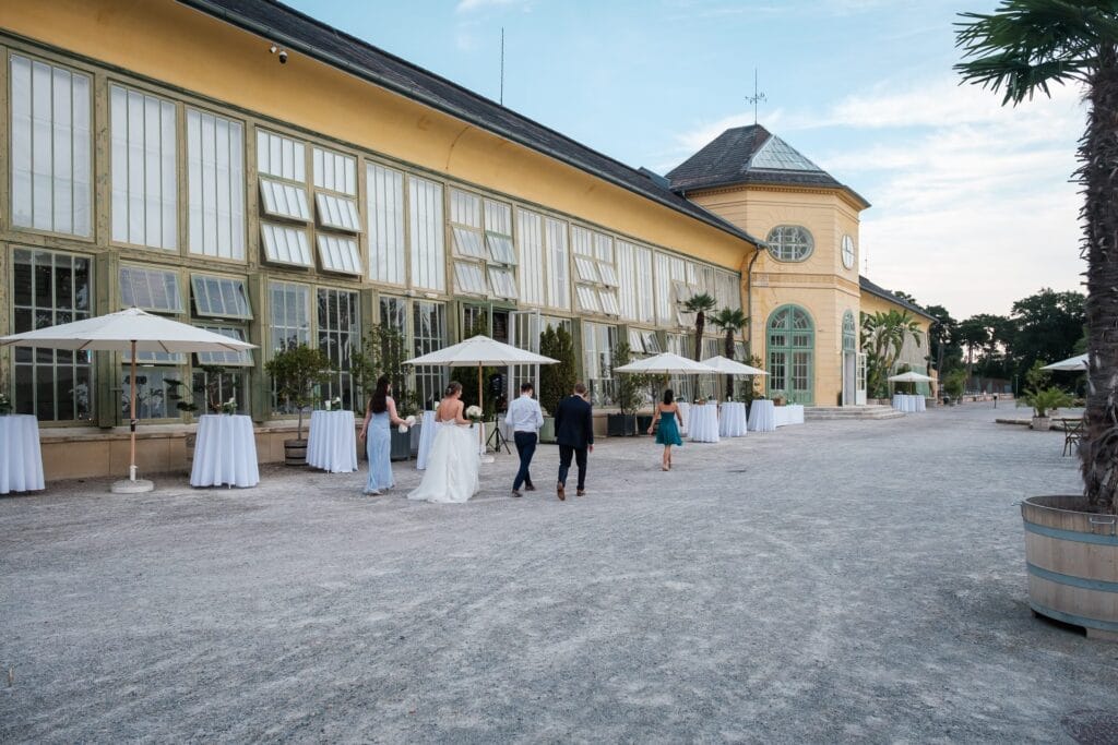 Hochzeitskrone Das Hochzeitspaar und die Gäste schlendern vor einem großen Gebäude mit Glasfenstern und gelben Wänden. In der Nähe säumen Tische mit weißen Sonnenschirmen den Kiesweg, perfekt für natürliche Hochzeitsfotos von einem talentierten Hochzeitsfotografen in Wien und Niederösterreich.