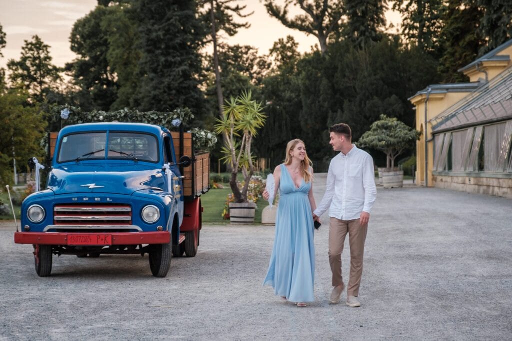 Hochzeitskrone Ein Paar schlendert auf einem Schotterweg neben einem blauen Oldtimer-Truck entlang, umgeben von üppigem Grün und einem gelben Gebäude – eine perfekte Szene für authentische Hochzeitsfotos.