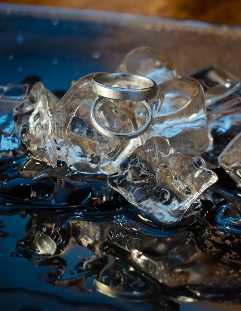 Hochzeitskrone Ein silberner Ring liegt auf schmelzenden Eiswürfeln und fängt echte Momente ein, indem er das Licht vor einem dunklen Hintergrund reflektiert.