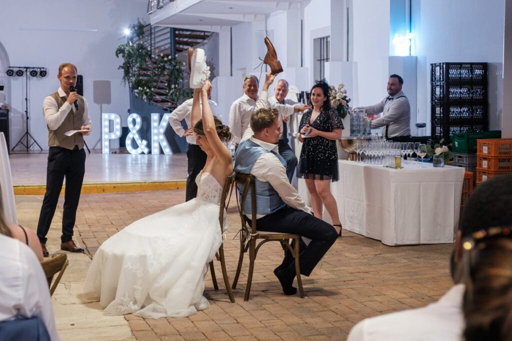 Hochzeitskrone Braut und Bräutigam spielen bei ihrer Hochzeitsfeier das Schuhspiel. Sie sitzen Rücken an Rücken und halten jeweils einen Schuh ihres Partners in der Hand. Diese echten Momente werden von einem Hochzeitsfotografen in Wien und Niederösterreich wunderschön festgehalten und verleihen ihren authentischen Hochzeitsfotos Charme.