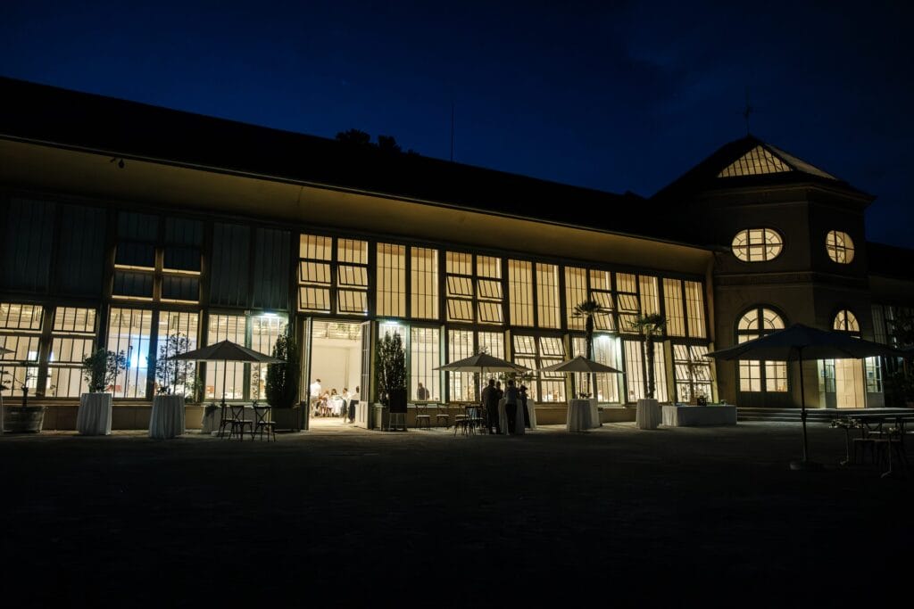 Hochzeitskrone Ein großes Gebäude mit hohen Fenstern wird nachts von innen beleuchtet und fängt echte Momente ein. Draußen sind mehrere Sonnenschirme und Tische aufgestellt, die den Charme natürlicher Hochzeitsfotos ausstrahlen. Durch die Fenster sieht man Menschen, die die Abendstimmung genießen.