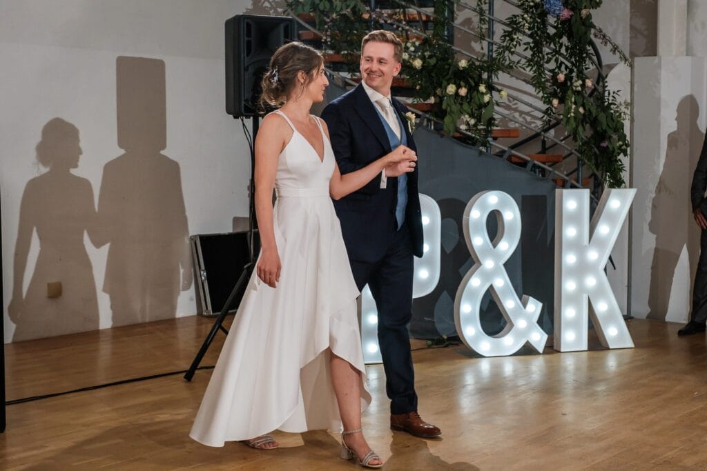 Hochzeitskrone Ein Paar tanzt anmutig in einem Raum, im Hintergrund leuchten die Leuchtbuchstaben „P & K“ sanft. Die Frau in einem fließenden weißen Kleid und der Mann in einem eleganten dunklen Anzug teilen diesen authentischen Moment, festgehalten von einem Hochzeitsfotografen in Wien und Niederösterreich.