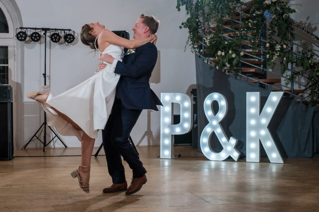 Hochzeitskrone Ein Paar tanzt fröhlich in einem Raum, im Hintergrund leuchten die Leuchtbuchstaben „P & K“. Er hebt sie lächelnd hoch und sie trägt ein weißes Kleid. So werden echte Momente eingefangen, die wirklich natürliche Hochzeitsfotos widerspiegeln.