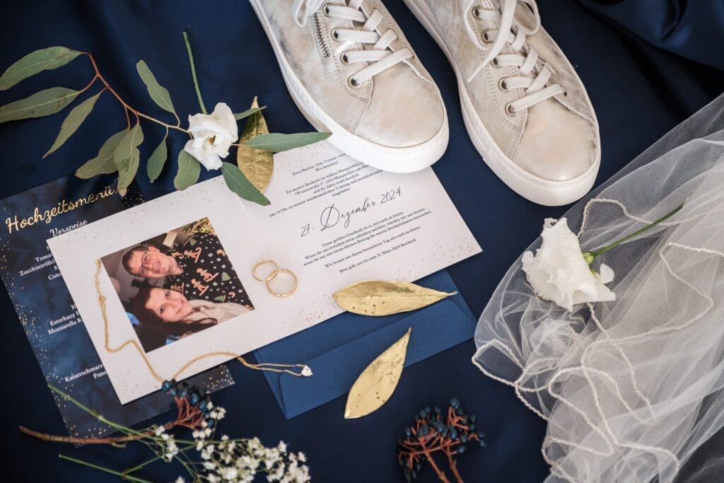 Hochzeitskrone Hochzeitseinladung mit natürlichen Hochzeitsfotos eines Paares, umgeben von goldenen Blättern, weißen Blumen, einem Schleier und silbernen Turnschuhen auf marineblauem Hintergrund.