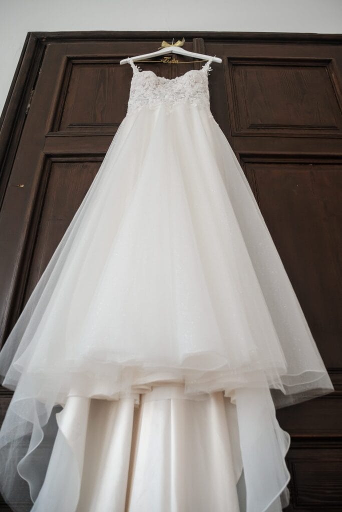 Hochzeitskrone Ein weißes Brautkleid mit Spitzenmieder und Tüllrock fängt bezaubernde Momente ein, wenn es elegant an einer Holztür hängt, perfekt für natürliche Hochzeitsfotos.