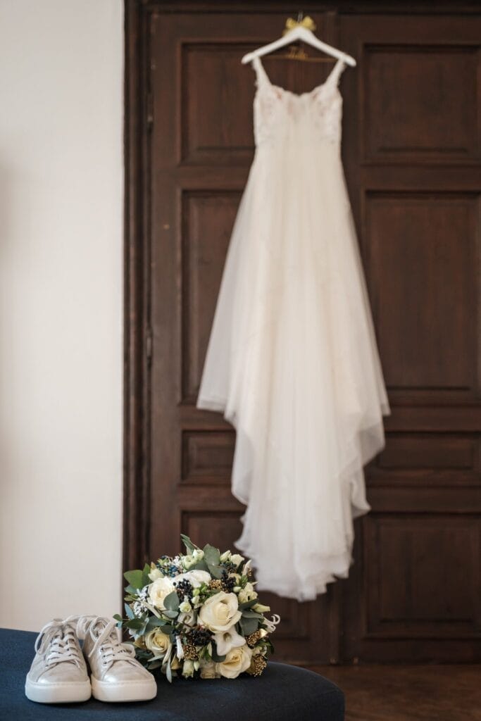 Hochzeitskrone Ein weißes Brautkleid hängt an einer Holztür und fängt bezaubernde Momente ein. Darunter vervollständigen ein Paar weiße Schuhe und ein Strauß mit weißen Rosen und Grünpflanzen diese idyllische Szene der Hochzeitsfotografie in Niederösterreich.