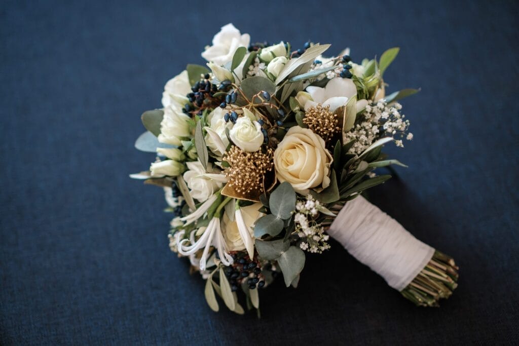 Hochzeitskrone Ein Strauß mit weißen Rosen, Grünpflanzen und kleinen weißen Blüten, umwickelt mit weißen Bändern, fängt bezaubernde Momente vor einem dunklen Hintergrund ein.